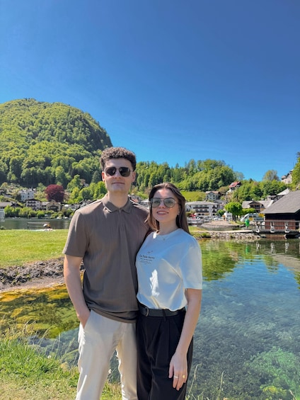 Two people standing by a clear lake with a hill and buildings in the background. 1