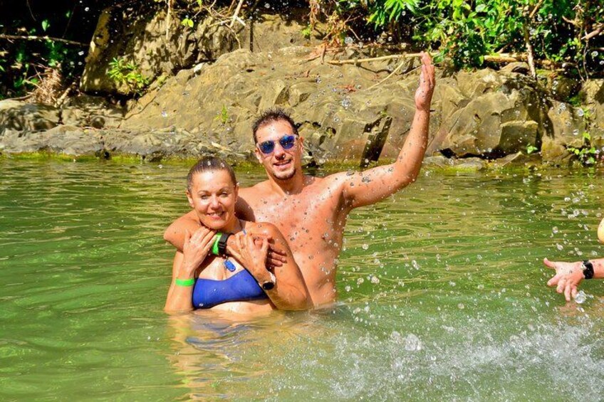 Deux personnes dans une étendue d’eau naturelle, l’une levant un bras, entourées de verdure et de rochers. 2