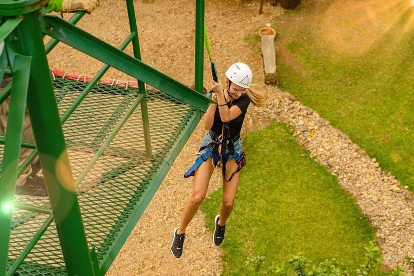 Una persona haciendo tirolesa con casco y arnés, una estructura de tirolesa metálica y un área con césped y un sendero. 2
