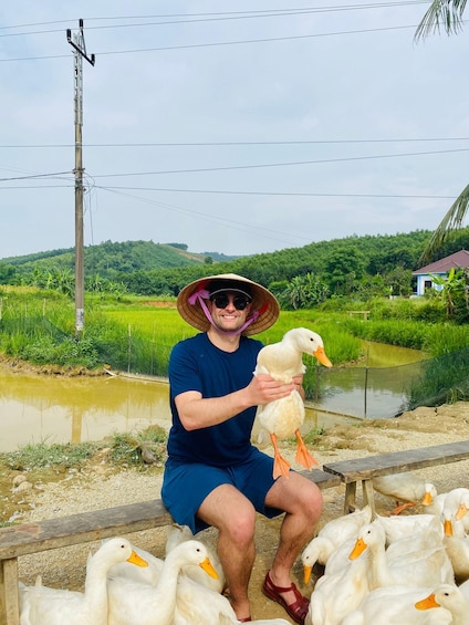 A person holding a duck, surrounded by other ducks, with a rural landscape and a water body in the background. 1