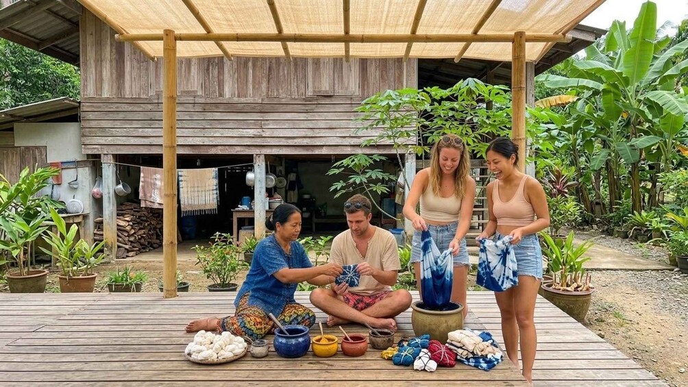 A group of people engaged in a craft activity outdoors, with a rustic wooden structure and lush greenery in the background. 2