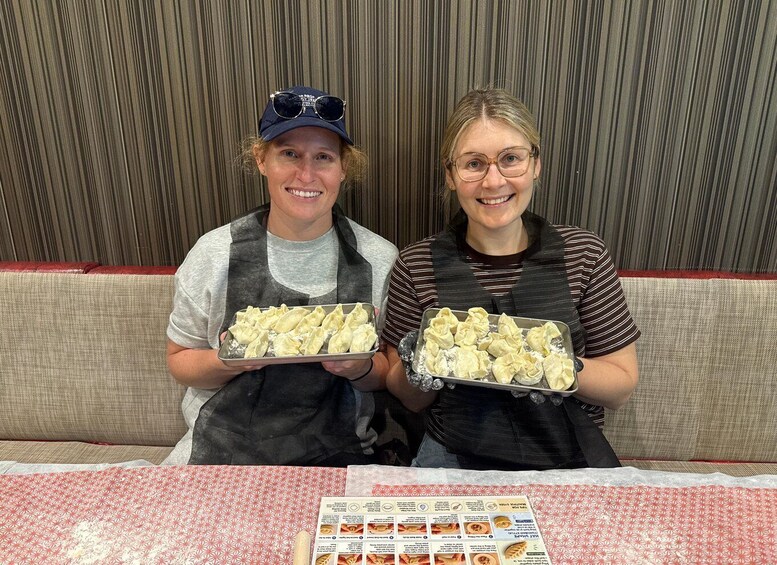 Two people holding trays of dumplings, seated at a table with a menu in front of them. 1