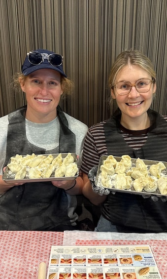 Two individuals holding trays of dumplings, with a recipe book on the table. 3