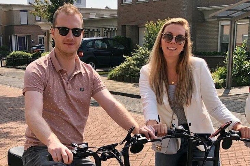 Two people on bicycles in an outdoor setting with buildings and a parked car in the background. 5