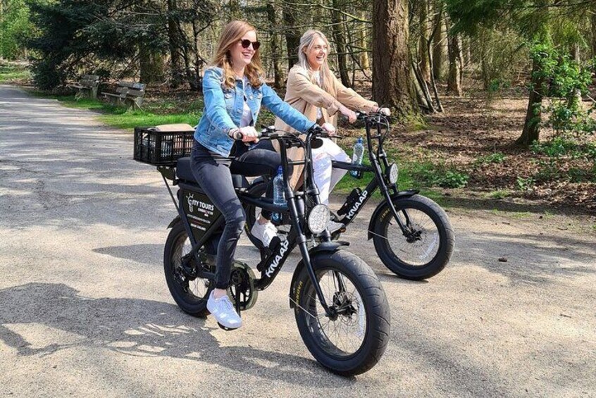Two people riding electric bicycles on a paved path in a wooded area. 1