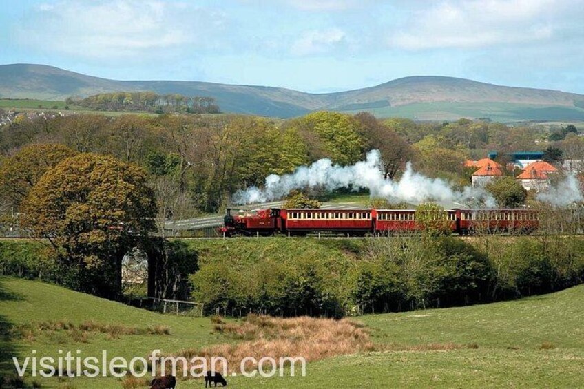 Un tren rojo con locomotoras a vapor viajando a través de un paisaje rural con campos verdes y colinas al fondo. 3