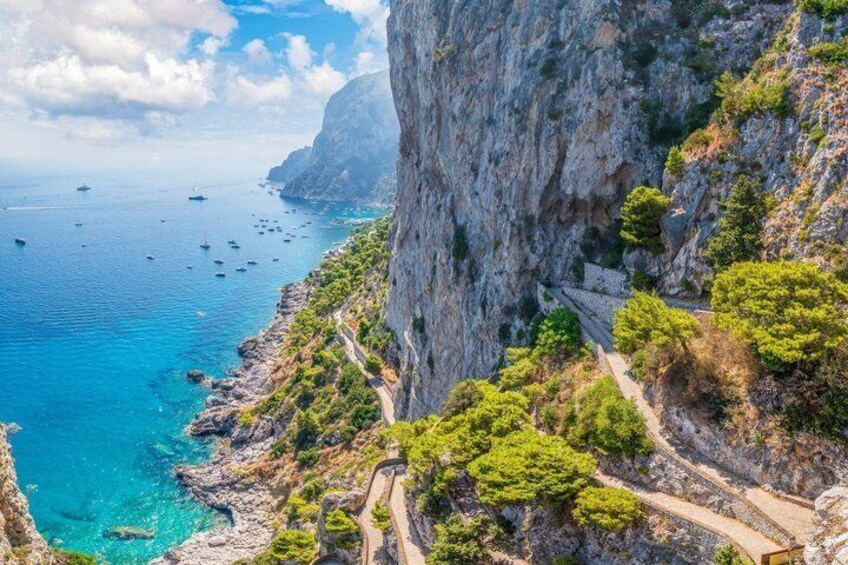 A coastal cliff with a narrow road, clear blue sea, and boats. 1
