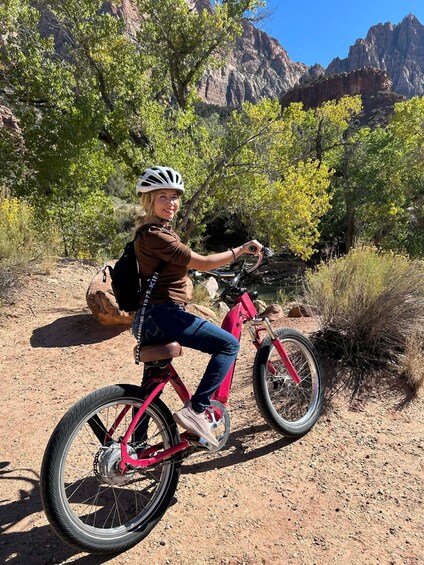 A person riding a pink bicycle on a dirt path with trees and rocky mountains in the background. 1