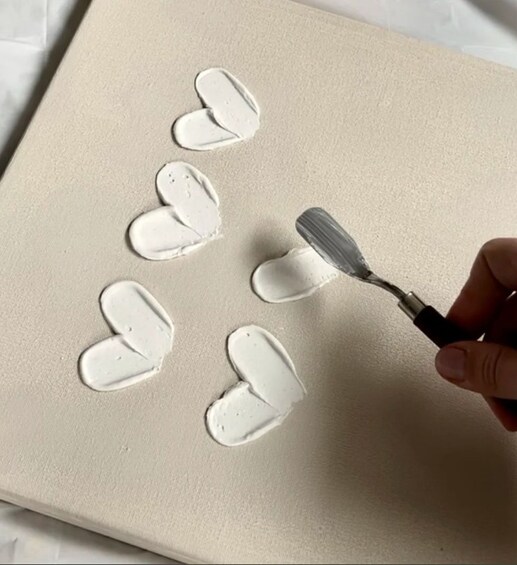 A hand applying white paint in heart shapes on a beige canvas. 1
