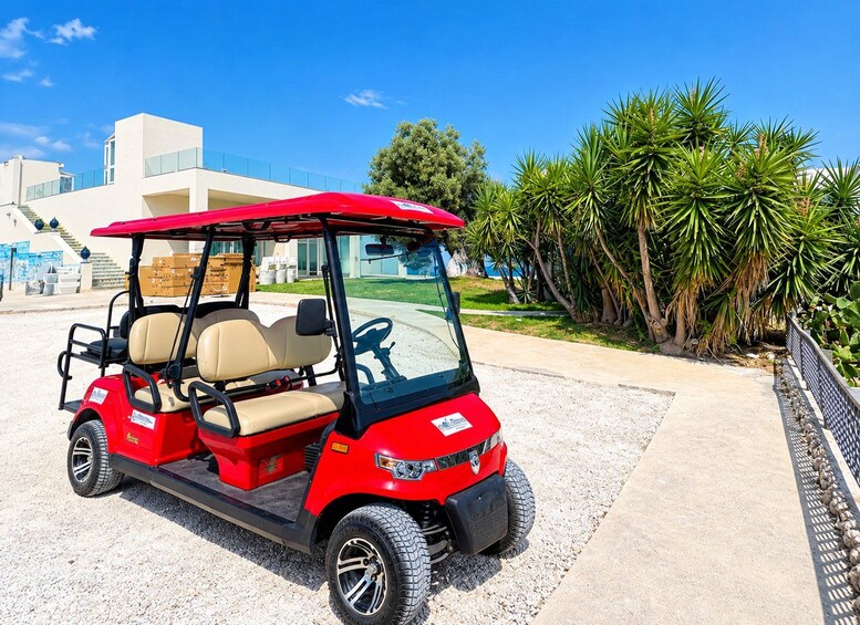 A red golf cart parked on a gravel path with a modern building and palm trees in the background. 1