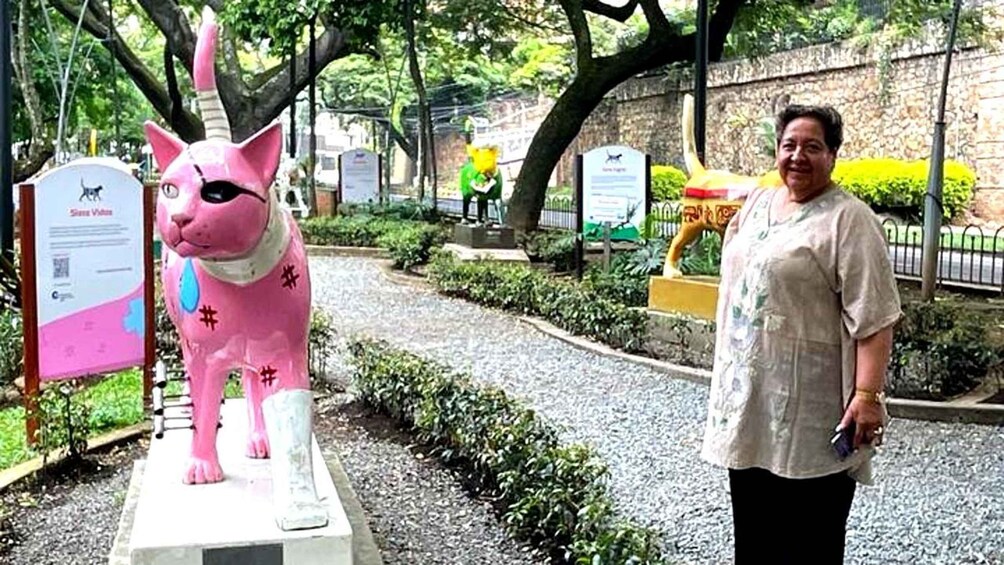 En person som står bredvid en rosa kattstaty i en trädgård med träd och en stenmur. 2
