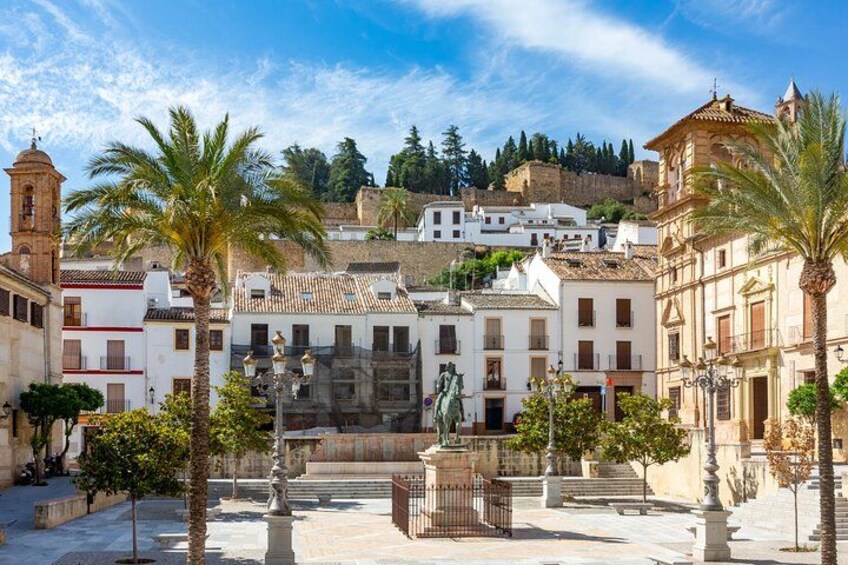 A historic town square with palm trees, a fountain, and buildings with white facades and red accents. 1