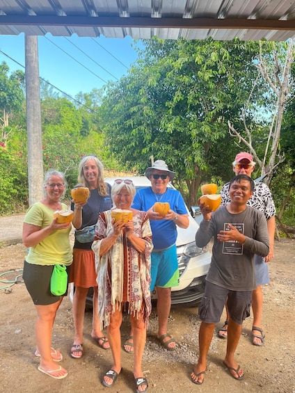 A group of people holding coconuts under a metal roof structure, with trees and a white vehicle in the background. 5