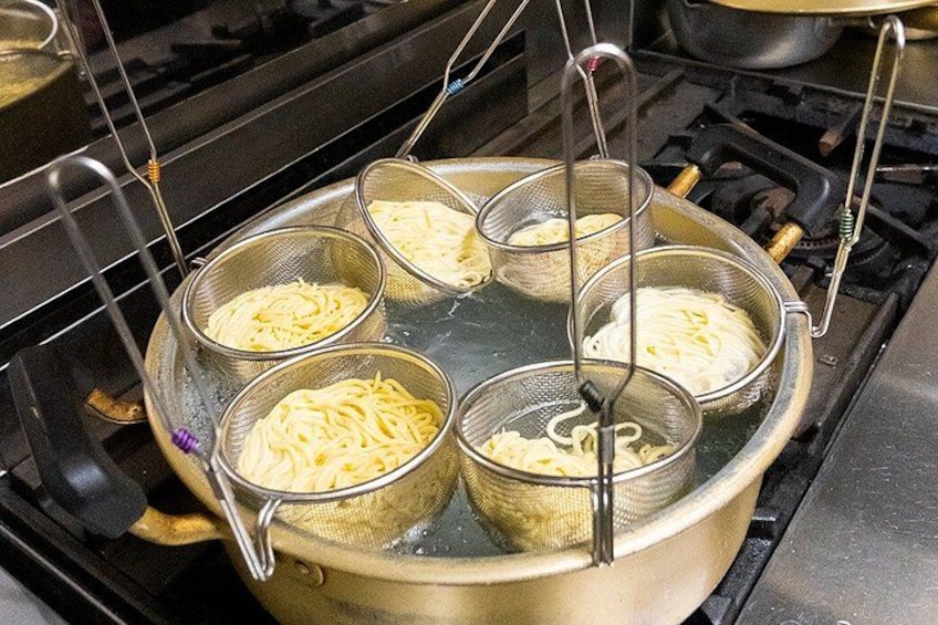A pot on a stove with four metal strainers containing noodles. 3