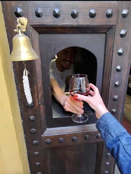 A person holding a wine glass, reaching through a wooden door with a bell and metal studs. 1