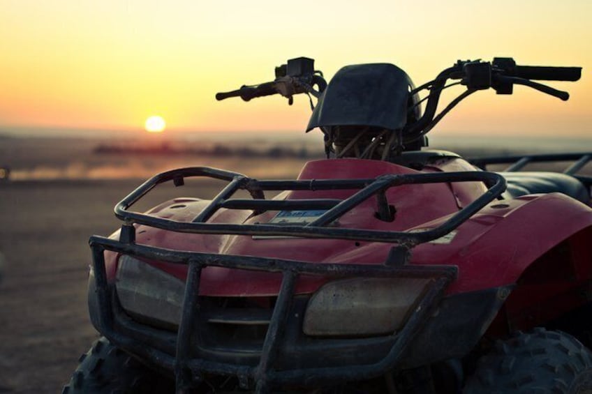 A red ATV with a front guard, parked on a dirt surface with a sunset in the background. 1