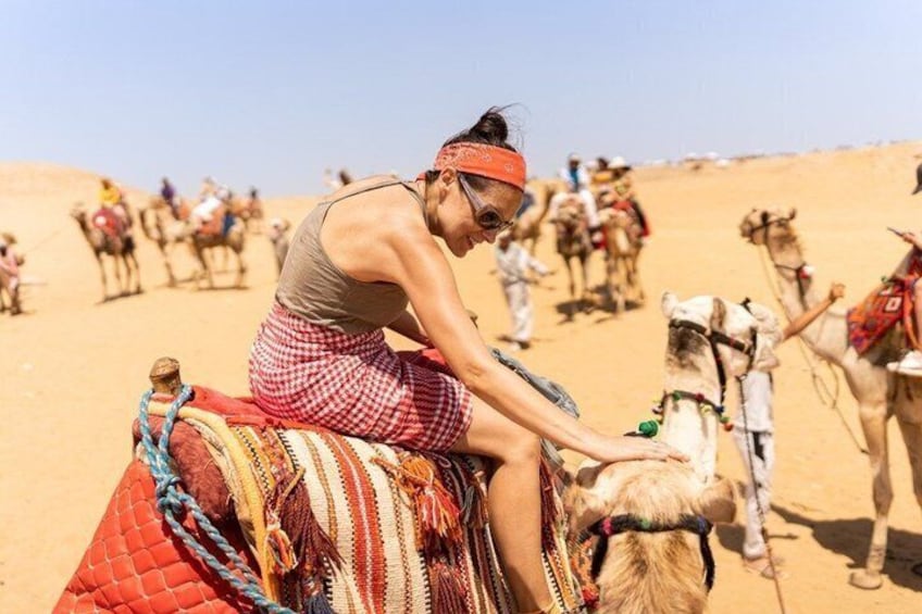 Una persona cavalca un cammello nel deserto, con altri cammelli e persone sullo sfondo. 3