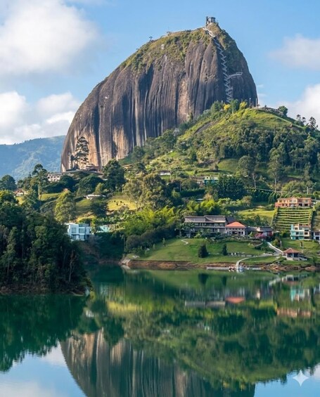 A large rock formation with a reflection in a lake, surrounded by greenery and buildings. 1