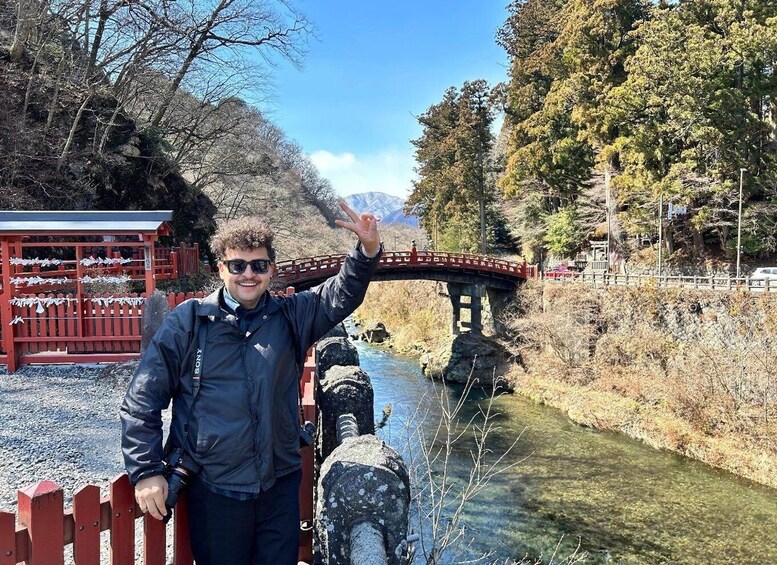 A person standing by a river with a stone bridge, trees, and a red structure in the background. 5