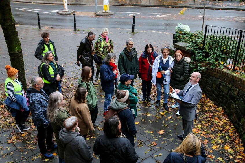 A group of people standing on a leaf-covered sidewalk, engaged in a discussion with a man holding papers. 5