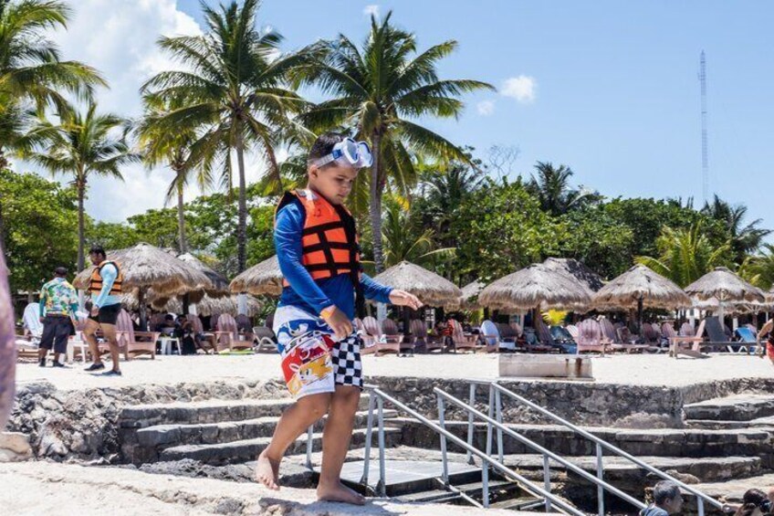 Una persona con chaleco salvavidas y equipo de snorkel en una playa arenosa con palmeras y construcciones de techo de paja. 3