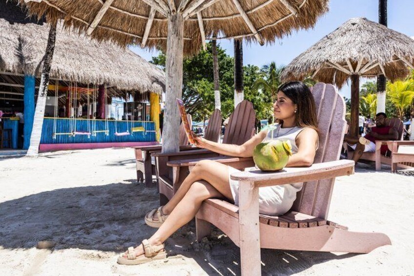 Una persona descansando en una silla de playa con una bebida de coco, bajo un toldo de paja. 4
