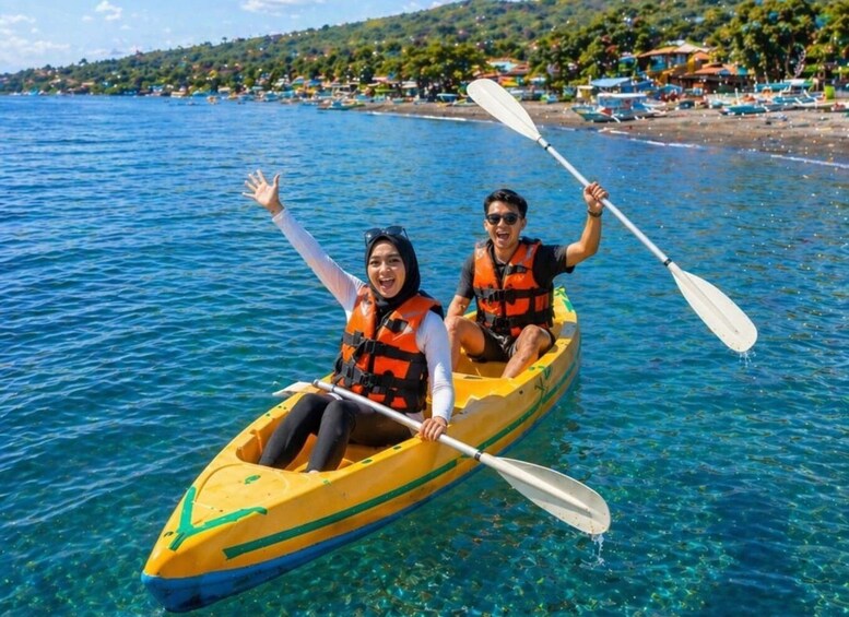 Two people kayaking on a clear blue sea with a beach and houses in the background. 3