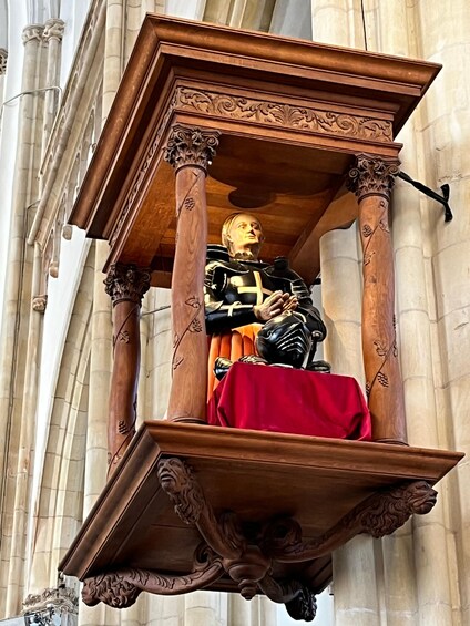 A statue of a knight in armor, mounted on a wooden structure with intricate carvings, set against a backdrop of classical architecture. 2