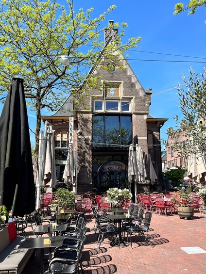 An outdoor cafe with red chairs and tables, a brick building with a large window, and a tree with green leaves. 3