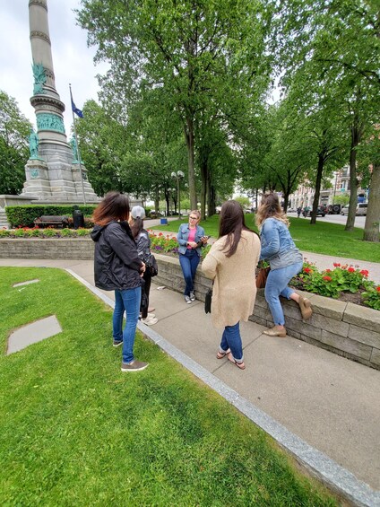 A group of people standing and conversing near a statue in a park with trees and a street in the background. 2