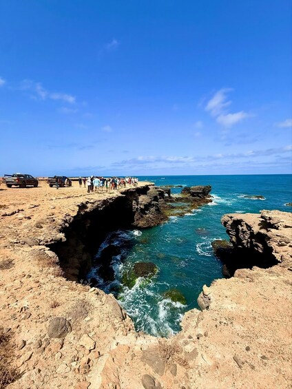 A rocky coastline with clear blue waters, a group of people on the shore, and a Toyota truck parked on the beach. 3