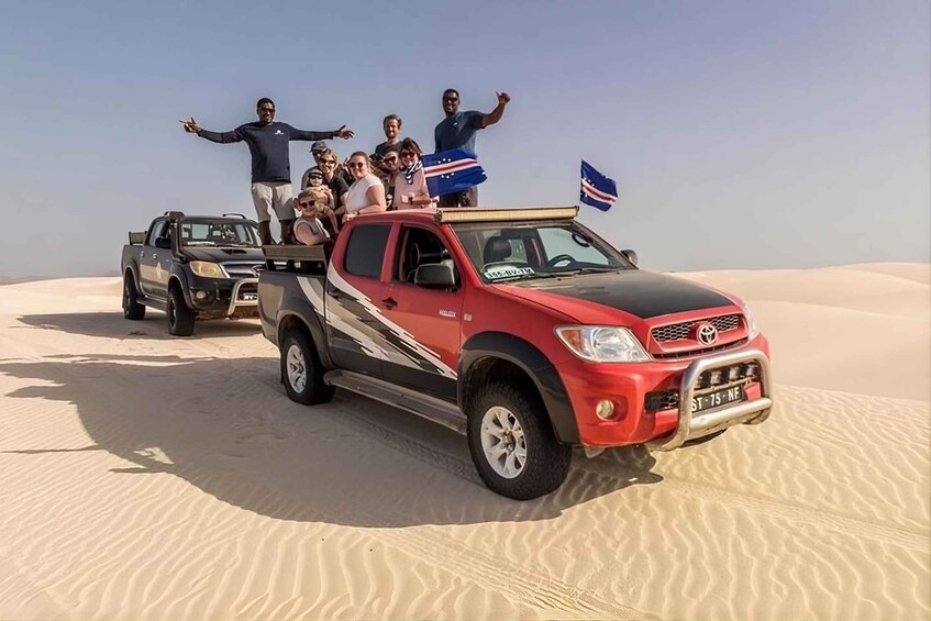 A red pickup truck with a group of people on a sand dune. 1