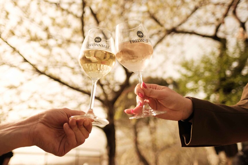 Two people holding wine glasses with 'FREDANIELE' labels, toasting in a natural setting with trees in the background. 2