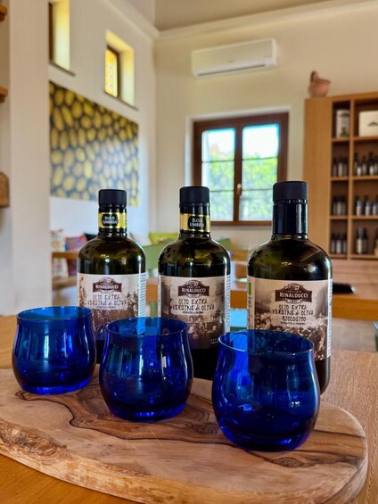 Three bottles of Rinalducci extra virgin olive oil and four blue glasses on a wooden board. 1