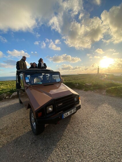 A brown off-road vehicle parked on a gravel path with a coastal landscape and a lighthouse in the background. 2