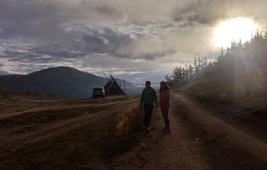 A dirt road with a van and a pile of sticks, surrounded by mountains and trees under a cloudy sky. 5
