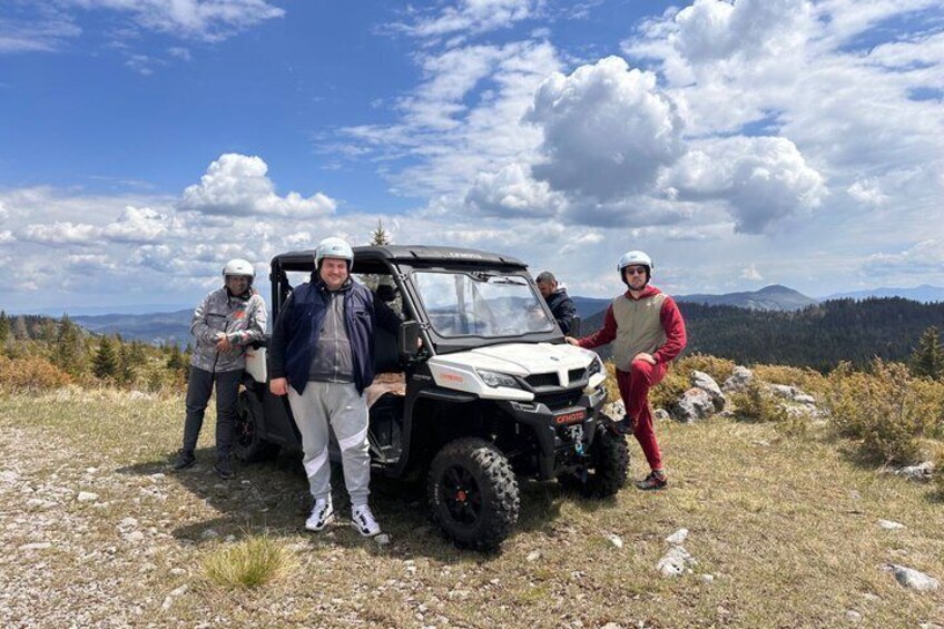 Eine Gruppe von Menschen steht neben einem weißen Geländewagen auf einer Wiese, im Hintergrund sind Berge zu sehen. 2