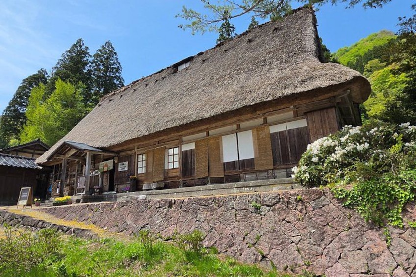 A traditional wooden building with a thatched roof, surrounded by greenery and a stone wall. 4