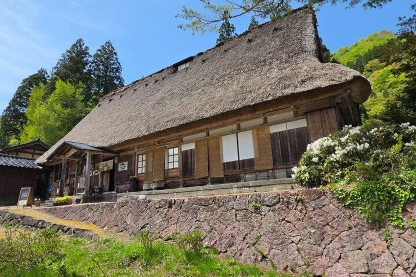 A traditional wooden building with a thatched roof, surrounded by greenery and a stone wall. 4