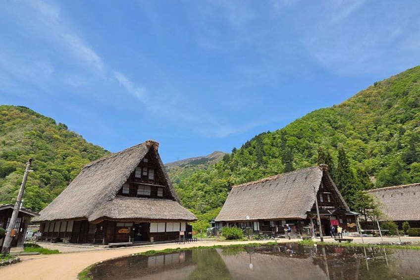 Traditional thatched-roof buildings, a calm pond, and lush green hills in the background. 5