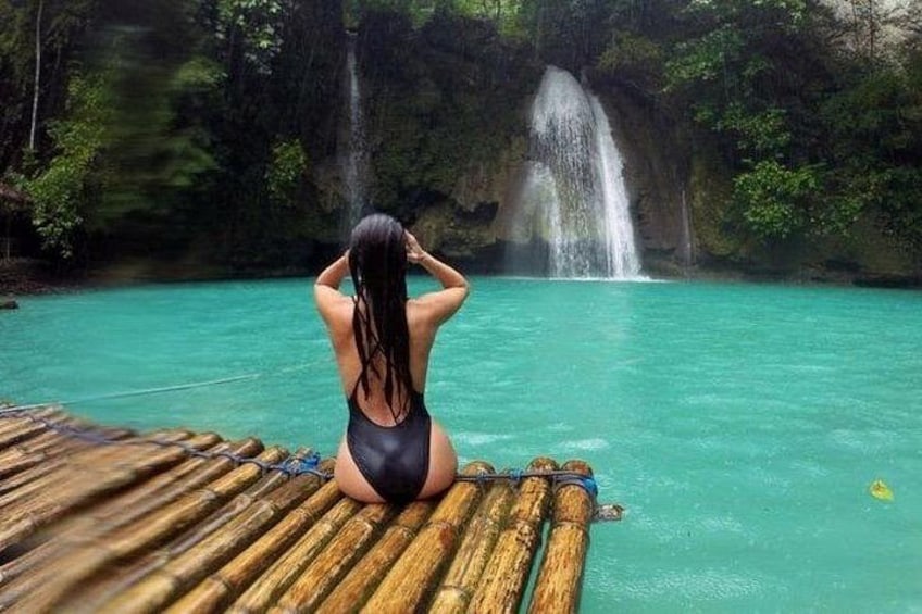 A person sitting on a wooden dock by a turquoise body of water, with a waterfall in the background. 3