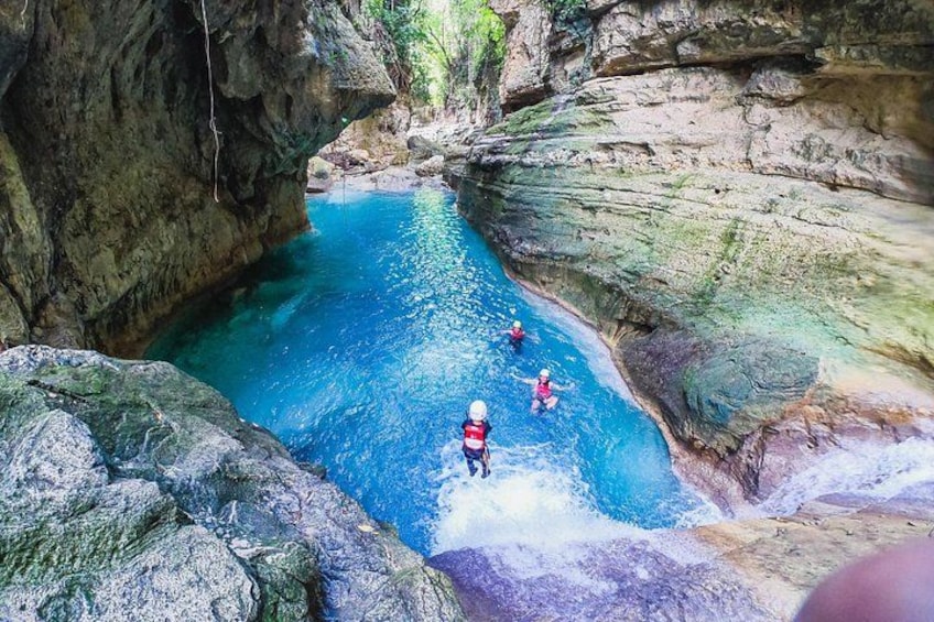 A canyon with a clear blue pool, rocky walls, and people swimming. 4