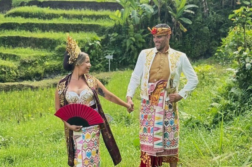 Zwei Personen in traditioneller Kleidung halten Händchen in einem üppigen, grünen Feld, im Hintergrund sanfte Hügel. 5