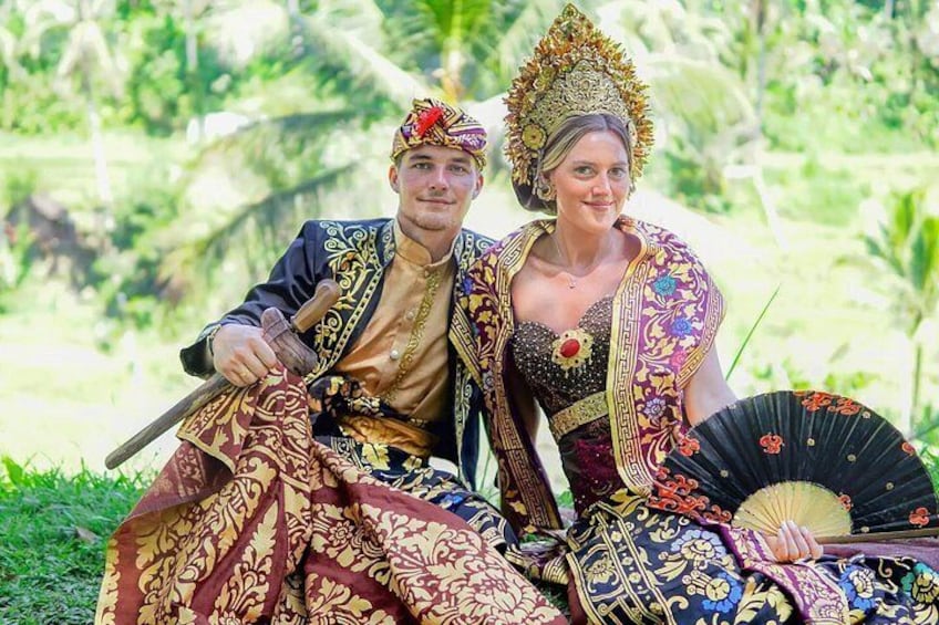 Ein Paar, gekleidet in traditioneller Tracht, sitzt draußen vor einer üppigen Grünanlage. 1
