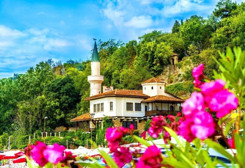 A white building with a tower, surrounded by lush greenery and pink flowers. 1