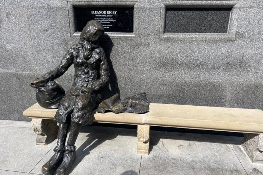 En bronze statue af en siddende figur på en bænk, med en plade med teksten 'Eleanor Rigby' i baggrunden. 4