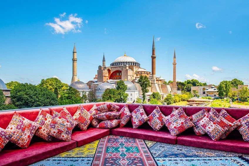 A view of a mosque with two minarets and a dome, a patterned carpet, and a red bench with cushions. 1
