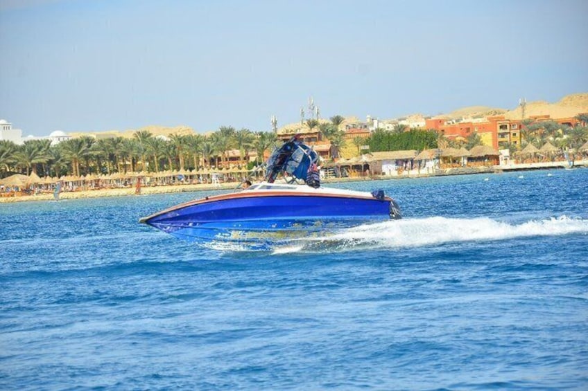 Een blauwe speedboot op het water, met een strand en gebouwen op de achtergrond. 2