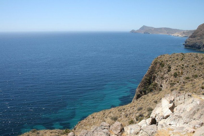 Eine Küstenlandschaft mit klarem, blauem Meer, felsigem Gelände und sanften Hügeln im Hintergrund. 2