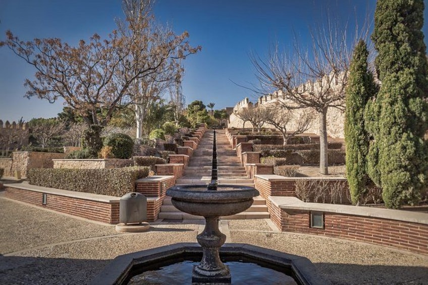 Ein Brunnen, ein Pflasterweg und ein Hügel mit Bäumen und einer Mauer im Hintergrund. 5